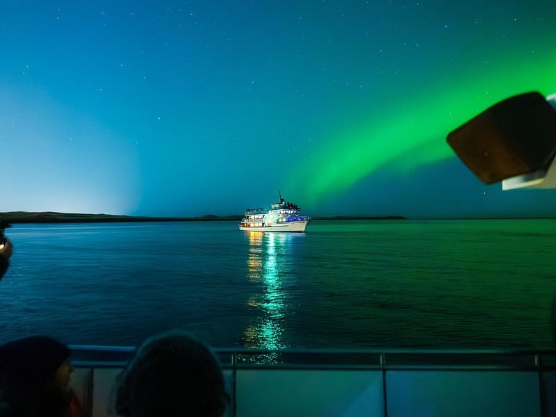 A lit ship on a calm bay under a green aurora, the rail of another boat in front
