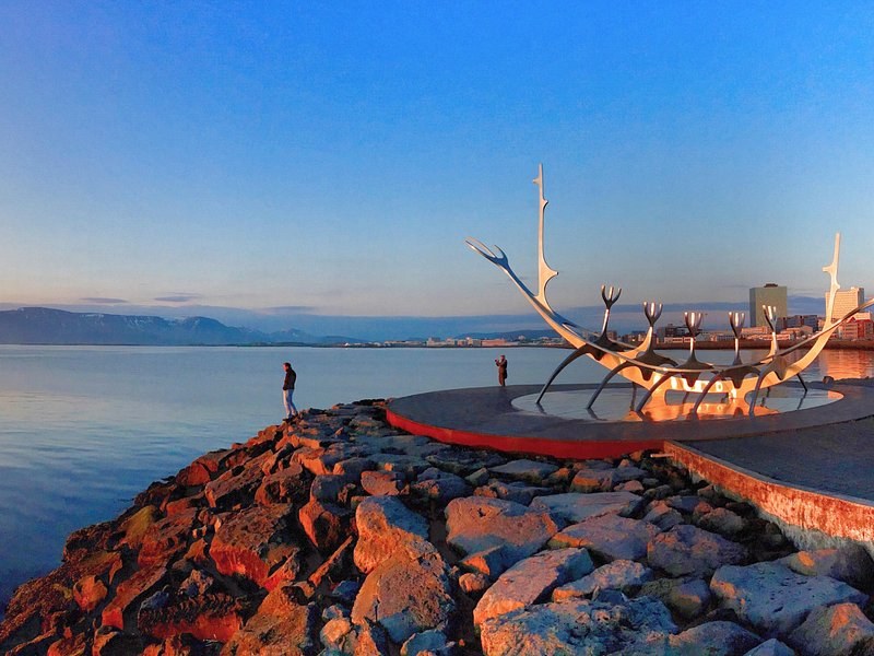 A steel ship-shaped sculpture on the city shore at sunset, the bay behind it