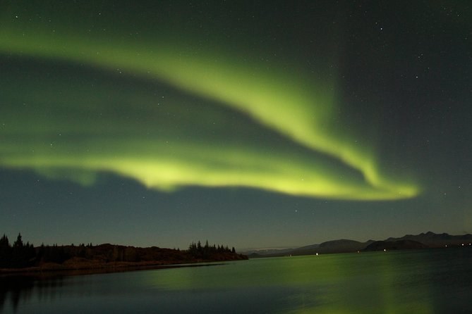 A pale green aurora arc over a lake and a line of dark trees