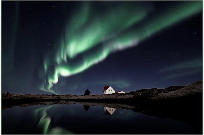 A white farmhouse beside a still pond, the aurora reflected in the water