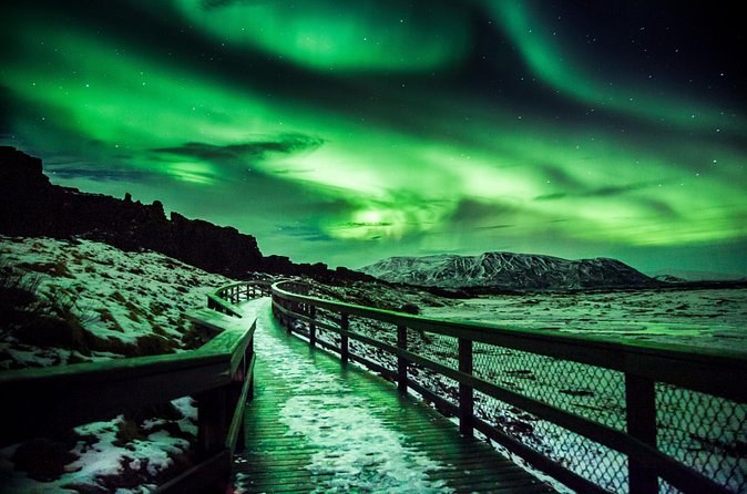 A railed boardwalk through snowy lava under a sky full of aurora