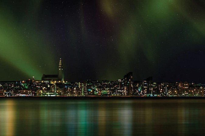 Green aurora over a city skyline and its church tower, lights reflected in the bay