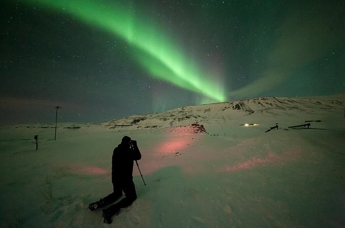 A photographer kneeling in the snow with a tripod under a green aurora