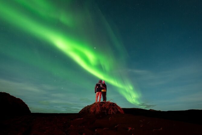 Two people standing on a rock outcrop under a sweeping green aurora